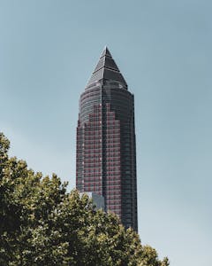 Modern skyscraper in Frankfurt under a clear blue sky, surrounded by greenery.