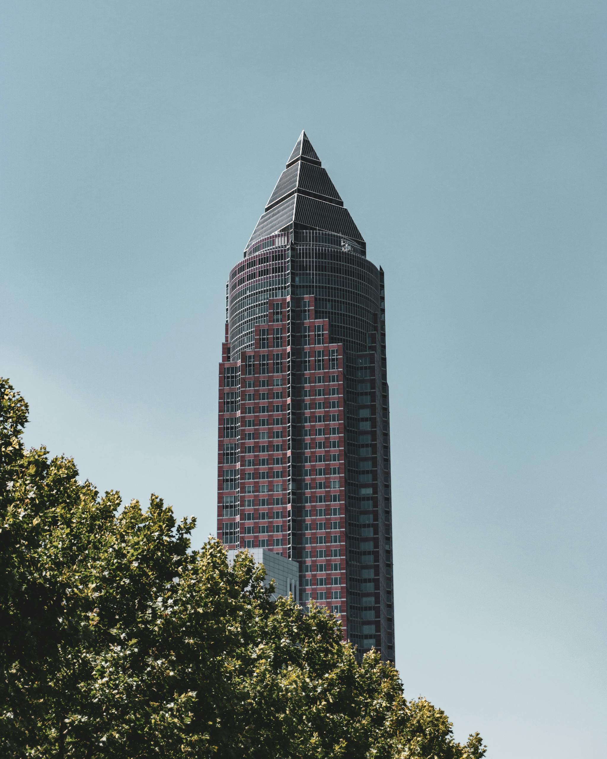 Modern skyscraper in Frankfurt under a clear blue sky, surrounded by greenery.