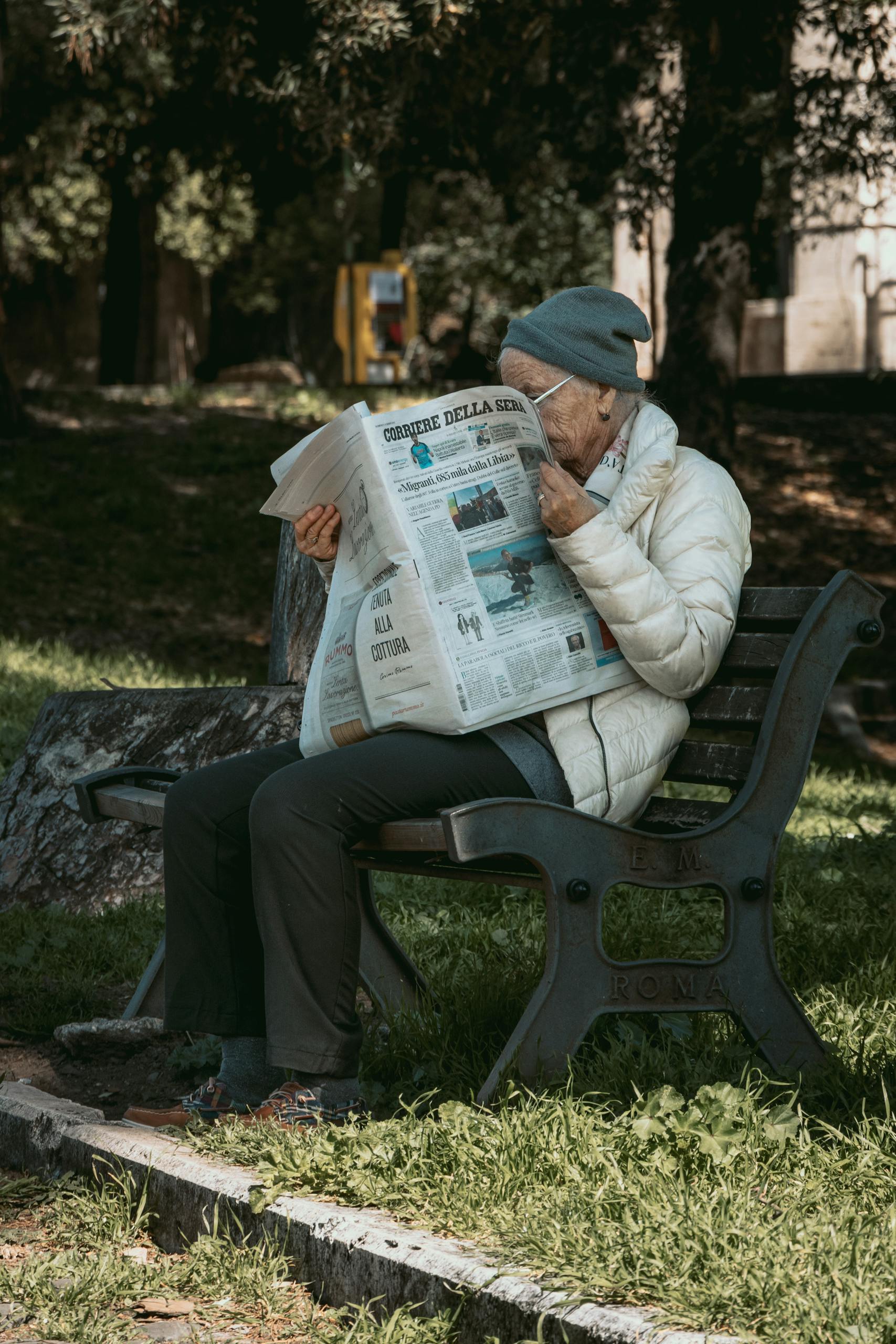 Senior man wearing winter attire reads Italian newspaper, outdoors in urban park.